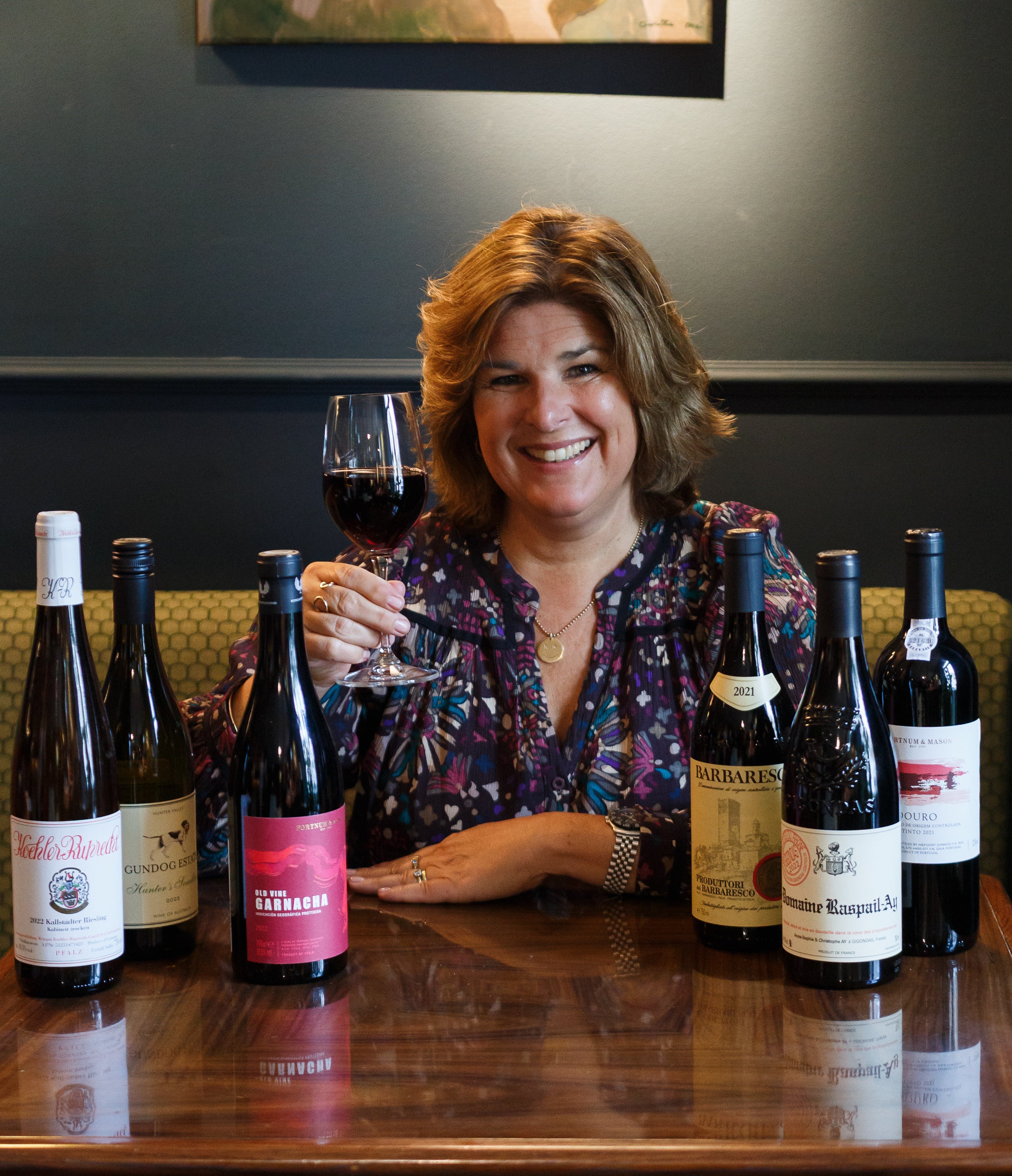 Helen sitting at a table holding a glass of red wine in her hand and smiling. All six bottles of wine in the autumn case are on the table in front of her.