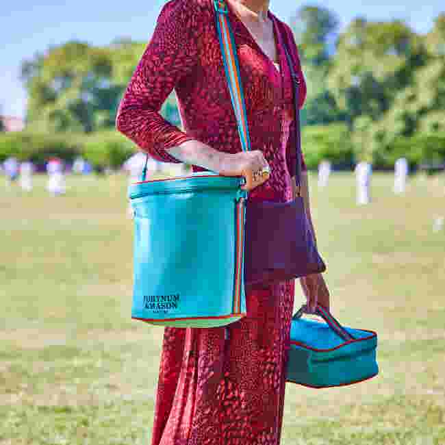 A woman in a red dress holds a blue Fortnum's picnic bag and lunch box while standing in a field.