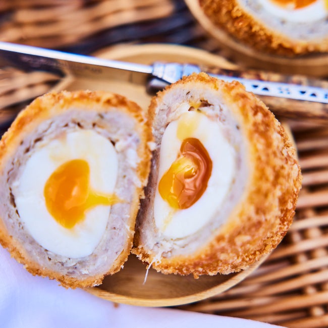 A scotch egg with a golden runny yolk is displayed in a bowl on a picnic basket